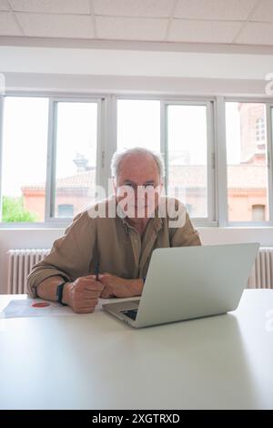 An elderly man sits at a bright office desk, focusing on work on his laptop He appears professional and engaged, embodying experience and dedication i Stock Photo