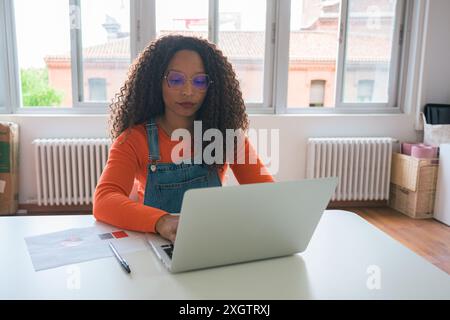 A young black woman focuses intently on her laptop in a sunlit home office setting Dressed casually in a denim jumpsuit and orange shirt, her curls fr Stock Photo