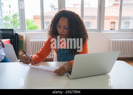 A focused businesswoman reviews documents and uses a laptop in a well-lit office environment, showcasing professionalism and productivity Stock Photo