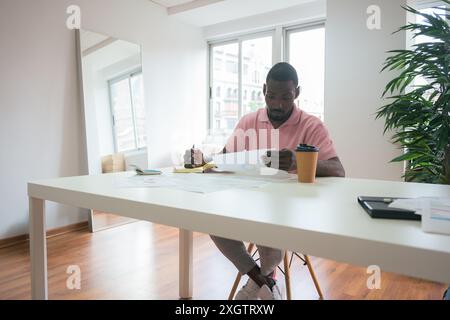 A professional man is intently focused on paperwork in a brightly lit, modern office, sitting at a white desk with coffee and a notebook by his side Stock Photo