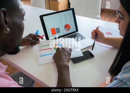 Two colleagues sit at a desk, deeply engaged in analyzing financial data displayed on a laptop and printed documents, discussing insights and strategi Stock Photo