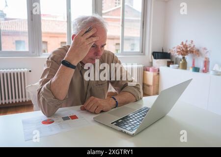 A distressed senior man examines financial charts on a table while sitting in a modern office, showing signs of stress as he holds his head with one h Stock Photo