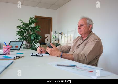 A focused senior businessman multitasks in a modern office space, holding a smartphone, and writing notes, surrounded by digital devices and office su Stock Photo