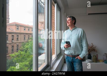 A middle-aged Caucasian man in a casual blue shirt and jeans stands by a sunny office window, holding a smartphone and looking thoughtfully outside at Stock Photo