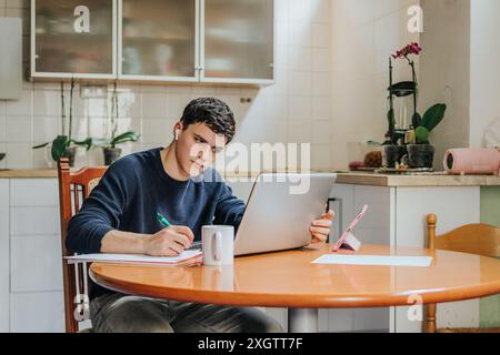 A focused young adult male studies from home, using a laptop and writing notes on a notebook at a kitchen table, with a coffee mug beside him and hous Stock Photo