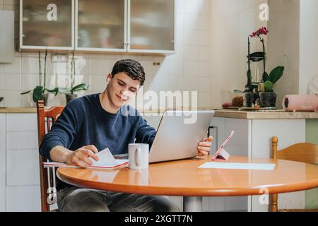 A young adult male is focused on his studies at a kitchen table, using a laptop and taking notes with a smartphone and notebook beside him. A serene h Stock Photo