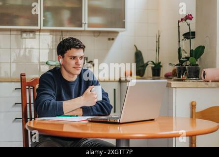 A young male sits at a kitchen table, engaging with a laptop while holding a coffee mug. A cozy home environment with plants and natural light surroun Stock Photo
