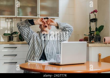 A young man in a robe sits at his home office table, visibly stressed, with his hands covering his eyes Stock Photo