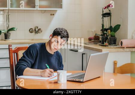 A young, focused man sits at a kitchen table, diligently working on his laptop while taking notes. Surrounded by a warm home environment, he personifi Stock Photo