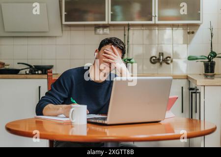 A young adult appears stressed and overwhelmed while sitting at a kitchen table with a laptop, papers, and a coffee mug, showing the challenges of rem Stock Photo