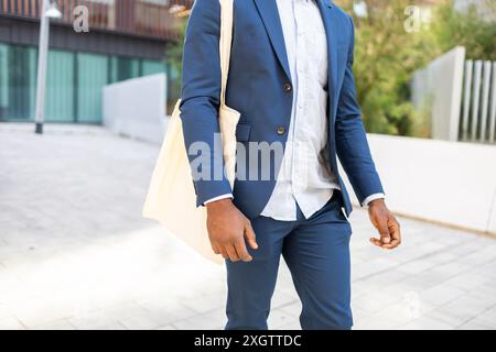 Cropped unrecognizable professional African American male walking confidently, dressed in a stylish blue suit and carrying a beige tote bag. He is loo Stock Photo