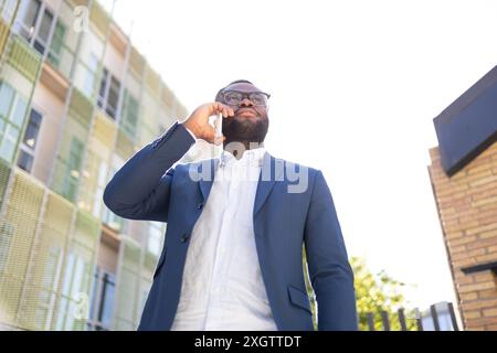 From below African American businessman engaged in a phone conversation, looking up thoughtfully in an urban setting, dressed in a stylish navy suit. Stock Photo