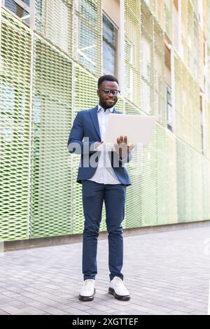 An African American businessman, wearing a smart blue suit and glasses, uses a laptop on a sunny day against a textured wall. He is smiling and lookin Stock Photo
