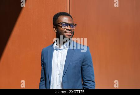 An African American man in a sophisticated blue blazer and glasses smiles comfortably. Positioned against a bold orange wall, he looks to the side, ad Stock Photo