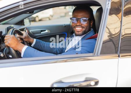 Cheerful African American businessman wearing glasses and a blue suit, driving a modern car and looking at the camera with a smile. Stock Photo