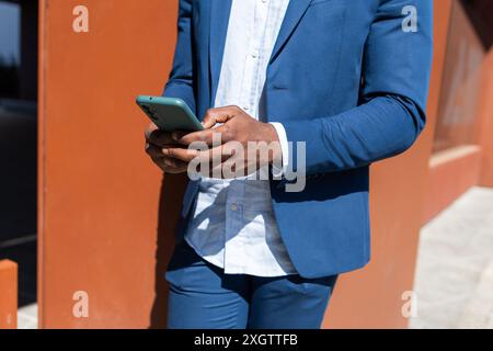 Cropped unrecognizable black business man in a blue suit using a smartphone against a orange wall Stock Photo