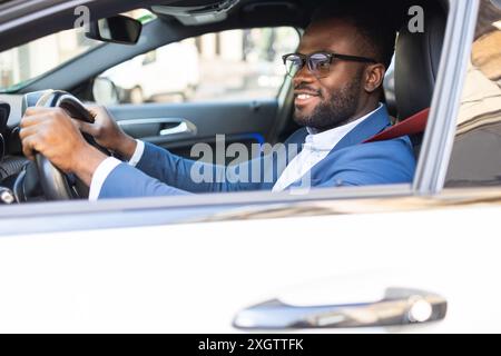 Confident African American businessman in blue suit driving his car, looking away thoughtfully while smiling slightly. Stock Photo