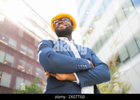 African American businessman in blue suit and yellow hard hat, standing outdoors with arms crossed and looking away confidently. Stock Photo