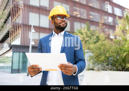 An African American businessman in a blue suit and yellow hard hat reviews documents while standing outside an urban office building. He looks away fr Stock Photo