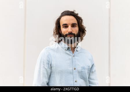 A portrait of a confident, young man with long hair and beard, dressed in a blue denim shirt, posing against a minimalist background He appears calm a Stock Photo
