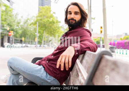A stylish young man with a beard rests on a bench in the Poblenou district, exuding a casual and comfortable lifestyle typical of the area Stock Photo