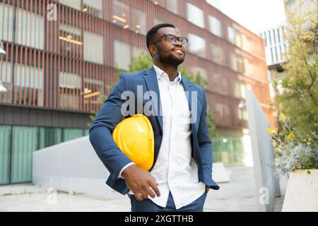 A confident African American businessman holding a yellow helmet walks near a contemporary office building, looking away from the camera. Stock Photo