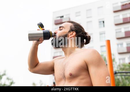 A bearded man drinks from a water bottle after exercising outdoors in Poblenou, Barcelona Apartment buildings serve as a backdrop, representing a blen Stock Photo