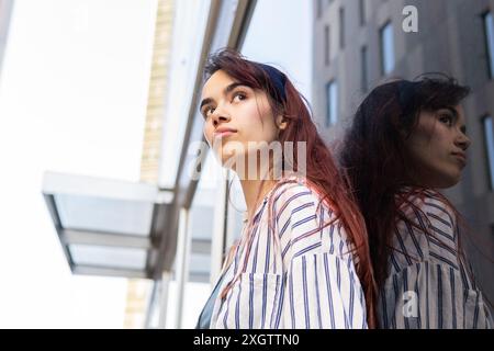 An introspective young woman with flowing hair looks upward, her image reflected on a glass building, expressing thoughtful energy in an urban environ Stock Photo