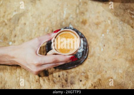 A close-up shot features a female hand with red nail polish holding a small espresso cup, presented on a rustically textured table surface Stock Photo