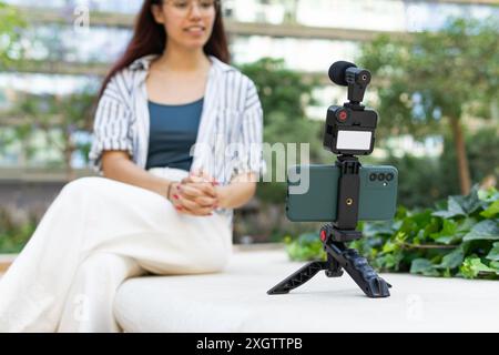 A young woman with redhead, wearing a striped shirt and white pants, is recording herself using a smartphone mounted on a tripod, with a blurred green Stock Photo