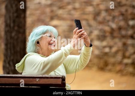 A cheerful elderly woman with blue hair taking selfie with her smartphone in a park, looking at screen with a smile. Stock Photo