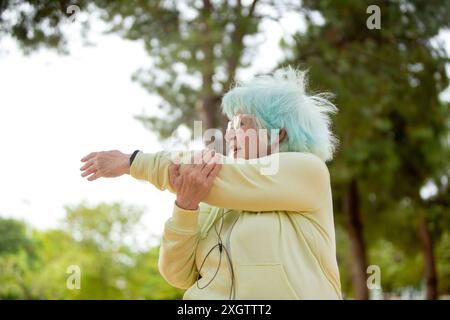 From below senior woman with blue hair is actively stretching her arms in a sunny park, looking away, dressed in a light yellow jacket. Stock Photo