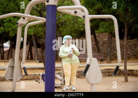 An elderly woman with blue hair enjoys a workout in a park, holding a water bottle and looking away. Stock Photo