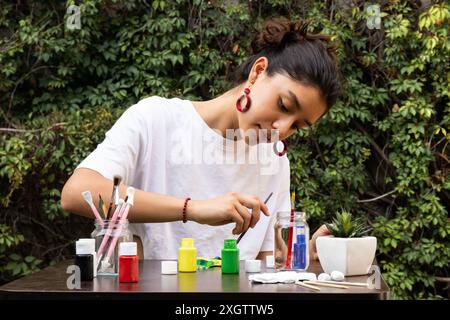 A focused young woman engages in the art of painting a clay pot, surrounded by a variety of paint pots and brushes on an outdoor table, set against a Stock Photo