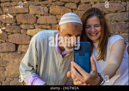 In Morocco, a smiling young woman shares a moment with an elderly man by taking a selfie together against a stone wall background The joy and intergen Stock Photo