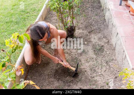 A woman kneels in her garden, using a hand hoe to prepare the soil for planting. She is barefoot and dressed in casual attire, focusing on garden main Stock Photo