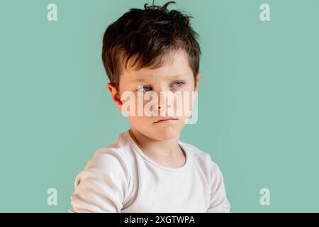 A young boy with dark hair and a solemn expression is photographed in a studio against a light teal background. He is wearing a white t-shirt and look Stock Photo