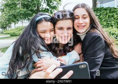 Three young female students sharing a cheerful moment together, huddled closely while taking a selfie in a lush park, vividly expressing friendship an Stock Photo