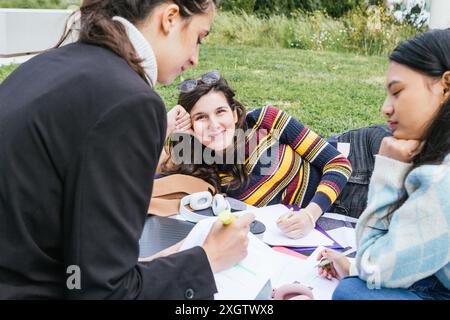Three university students gather outdoors on the grass with notebooks and pens, studying and enjoying a sunny day together. They display gestures of f Stock Photo