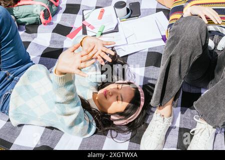 Three friends enjoying a sunny day outdoors while surrounded by study materials. One student is lying down, shielding her eyes, highlighting a relaxed Stock Photo