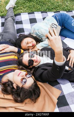 Three young students lounge on a checkered blanket in the park, sharing laughter and capturing memories with a smartphone Stock Photo