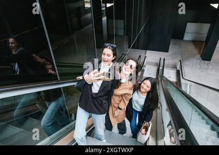 Three enthusiastic young students laugh and capture a selfie while riding down an escalator in a modern urban setting, enjoying their day out together Stock Photo