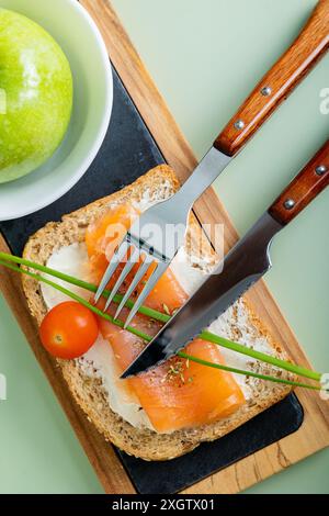 Top view of a nutritious breakfast featuring salmon on whole grain bread, garnished with fresh chives and cherry tomato, alongside a green apple. Stock Photo