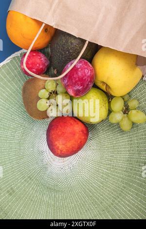 Top view of a vibrant selection of fresh fruits spilling from a paper bag onto a textured green mat. Includes oranges, avocados, apples, kiwis, and gr Stock Photo