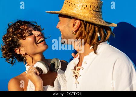 A joyful Caucasian woman and a Caucasian man with dreadlocks laugh together under a vivid blue sky. Both are dressed in summer clothes, looking at eac Stock Photo