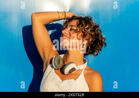 A joyful Caucasian woman with curly hair smiles while listening to music through white headphones, standing against a vibrant blue background Stock Photo