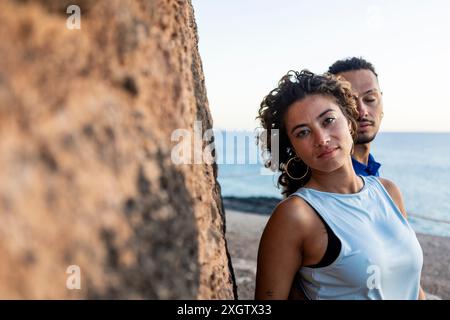 A Caucasian woman in focus with curly hair and large earrings gazes directly at the camera, while a man with dreadlocks stands behind her, near a rock Stock Photo