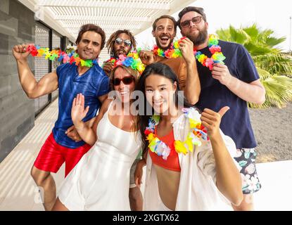 A vibrant group of multiracial friends celebrate summer, wearing colorful leis and festive clothing. They are smiling and waving, enjoying the sunny o Stock Photo