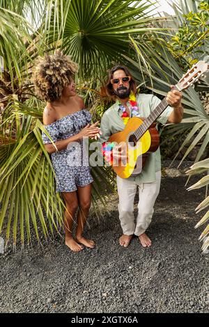 A joyful Hispanic male playing guitar accompanied by an African American female with curly hair floral accessories, set against lush tropical foliage. Stock Photo
