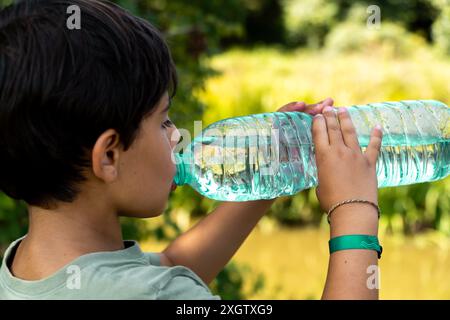 Side view of a boy quenches his thirst by drinking water from a large bottle while on a nature walk, highlighting a moment of refreshment in a lush gr Stock Photo